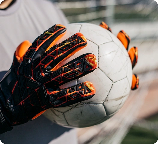 Luva de guarda-redes a segurar firmemente uma bola de futebol, mostrando a textura antiderrapante e detalhes de design em laranja e preto.