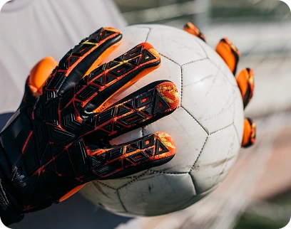 Luva de guarda-redes a segurar firmemente uma bola de futebol, mostrando a textura antiderrapante e detalhes de design em laranja e preto.