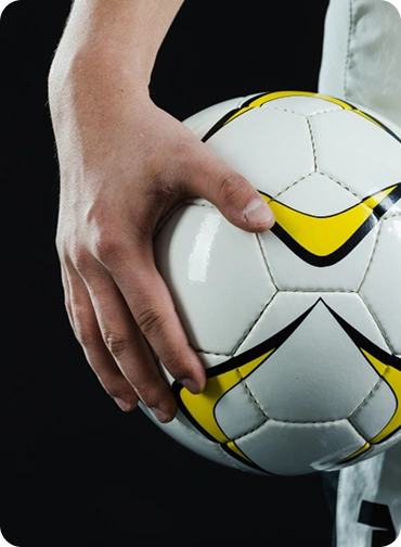 Primeiro plano de uma mão a segurar uma bola de futebol branca com detalhes amarelos, sobre um fundo escuro.