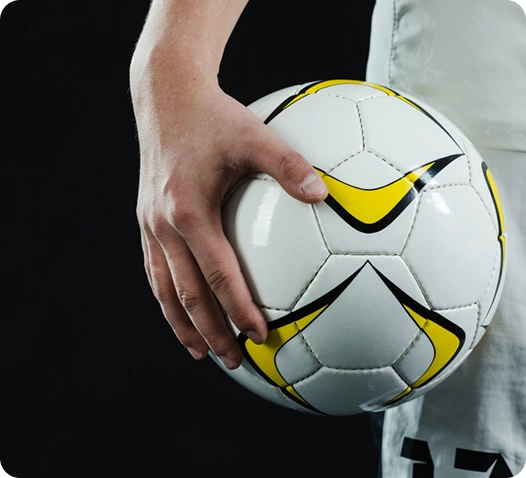 Primeiro plano de uma mão a segurar uma bola de futebol branca com detalhes amarelos, sobre um fundo escuro.